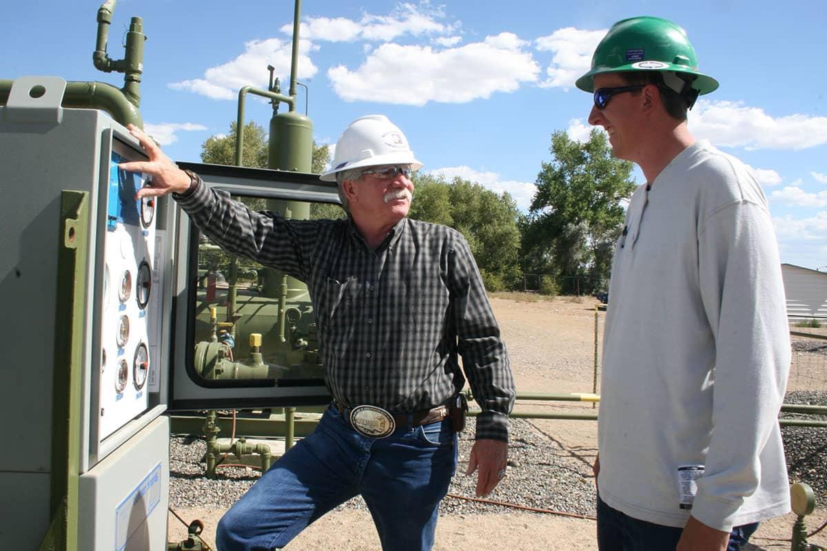 Two individuals in hard hats look at each other while standing next to a natural gas meter